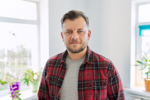 Ein Mann mittleren Alters mit kurzem Bart und kariertem Hemd steht entspannt in einem hellen Raum mit Pflanzen im Hintergrund. Er ist Patient mit Migräne.